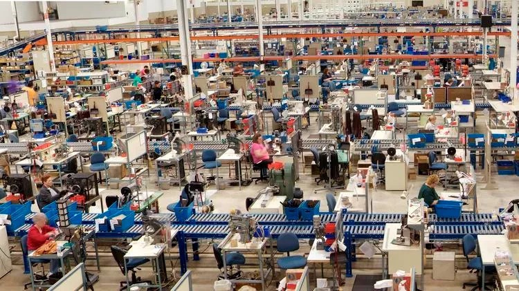 Large manufacturing floor with rows of workstations, conveyor belts, and assembly equipment, where multiple workers are seated or standing at stations assembling products under bright overhead lighting.