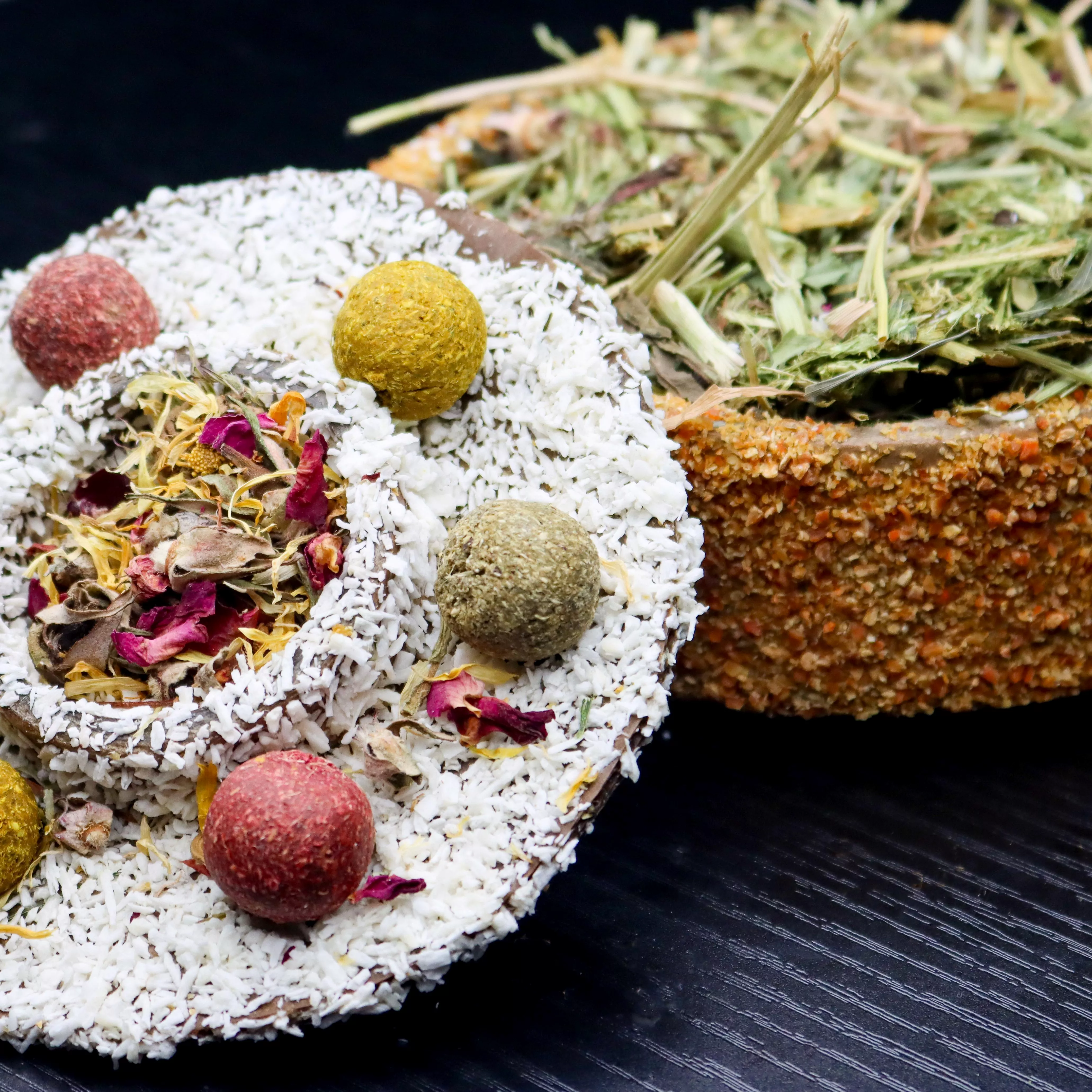 Close-up of an opened small animal celebration cake. The removable lid is topped with coconut flakes, dried flower petals, and colorful herb and vegetable treat balls. The cake base is coated in dried carrot pieces and filled with hay and herbal forage.