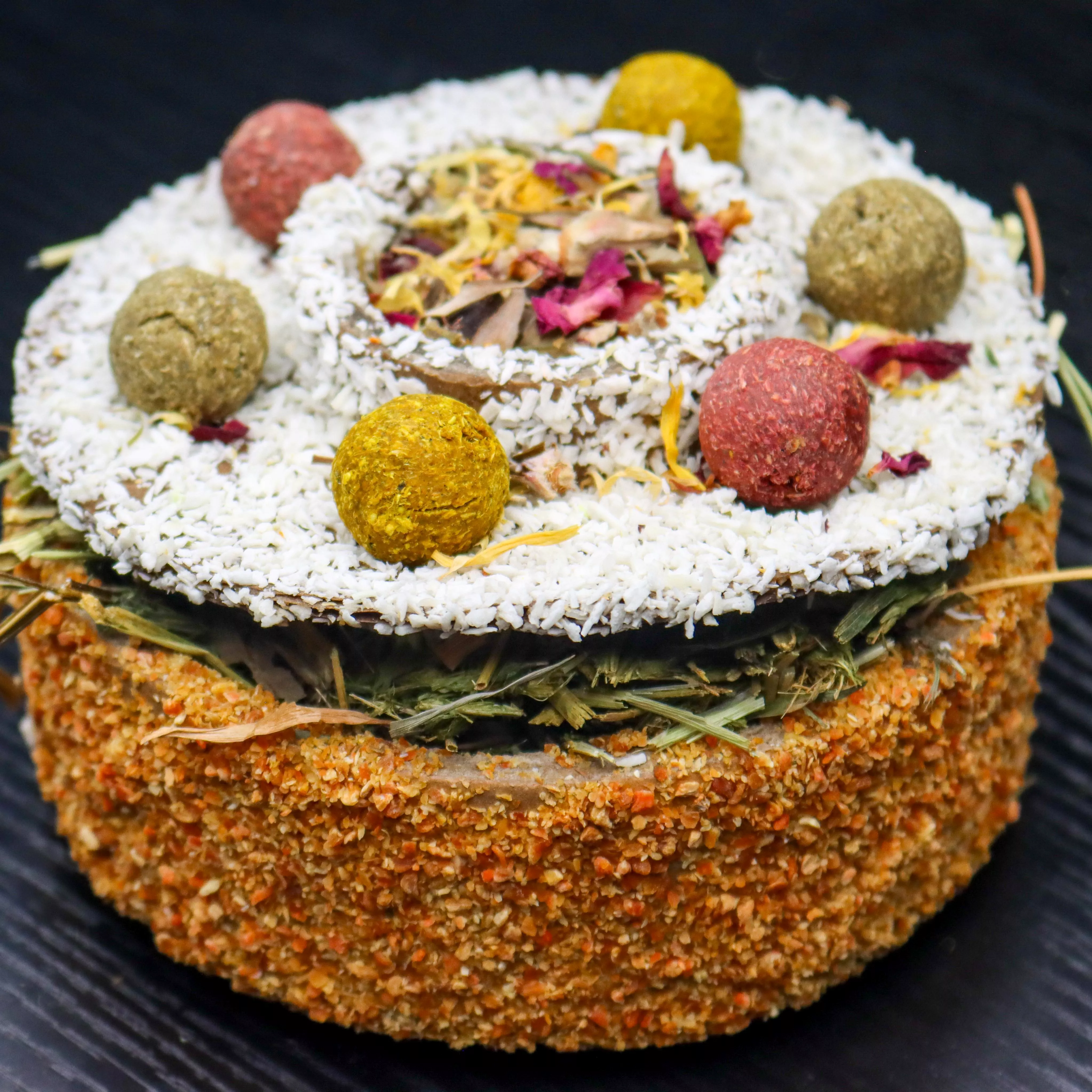 Close-up of a round small animal celebration cake coated in dried carrot pieces and topped with coconut flakes, colorful herb and vegetable treat balls, and a center section filled with dried flowers and forage. The cake is partially open, revealing a layer of hay and herbal forage inside.