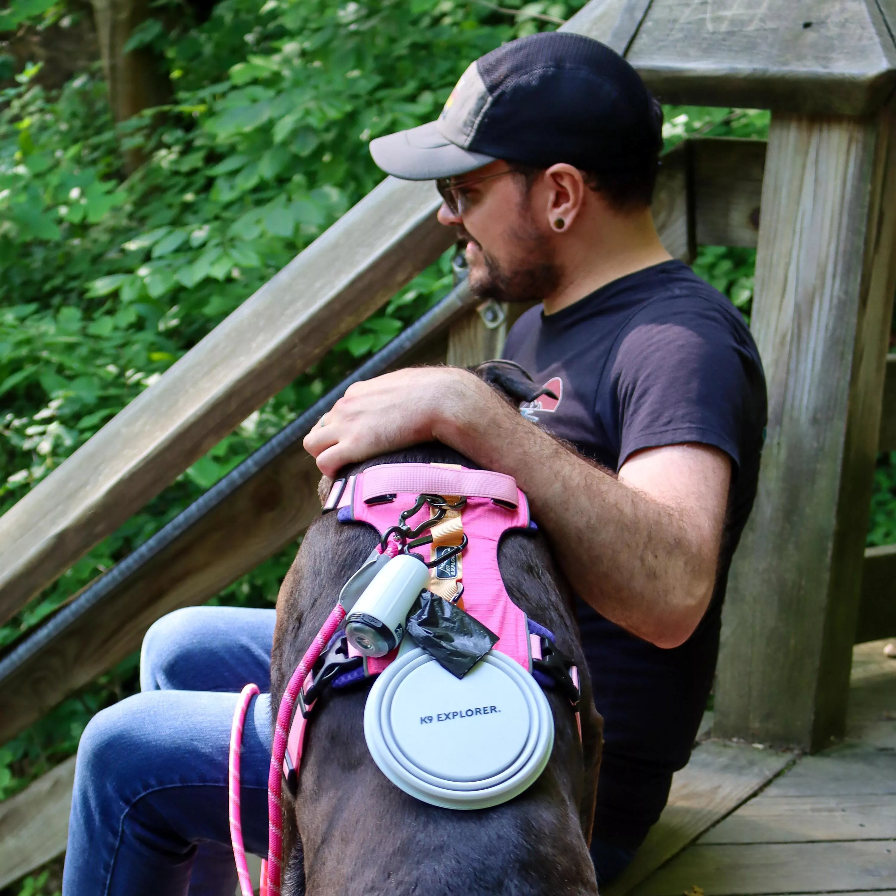 A dog wearing a pink harness is outdoors on a wooden walkway. A white K9 Explorer waste bag dispenser with an integrated light is clipped to a fabric loop on the harness using a black plastic hook. A round collapsible bowl in light gray is attached near the dispenser by a fabric loop. A pink braided rope leash is connected to the harness. The background includes green foliage and wooden support rails.