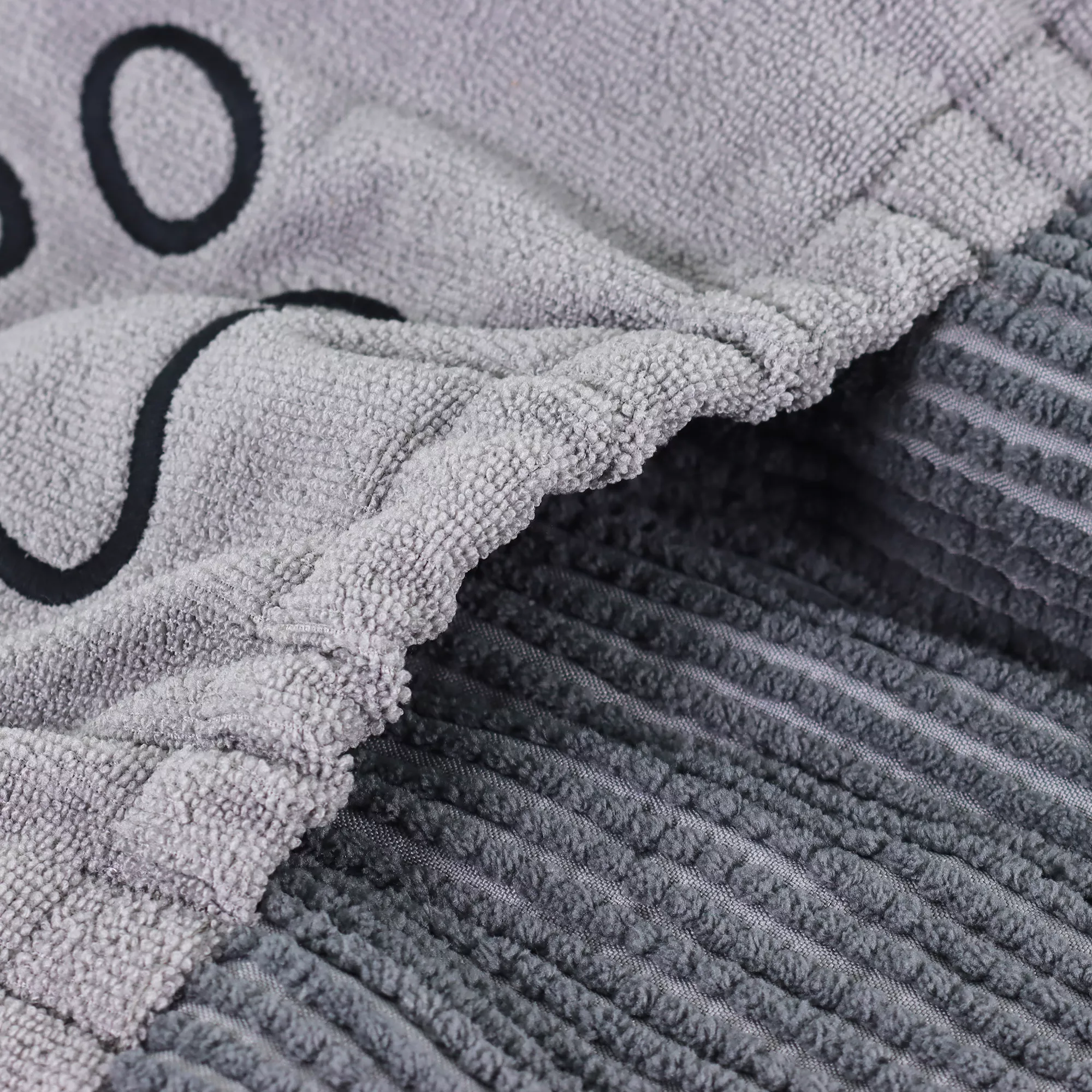 A close‑up view of the shammy towel shows the ribbed gray fabric of the pocket section. The pocket edge is slightly gathered with visible stitching. Part of an embroidered paw‑print design in black thread appears near the upper area of the fabric. The lower portion of the image shows the towel’s darker gray textured microfiber chenille surface.