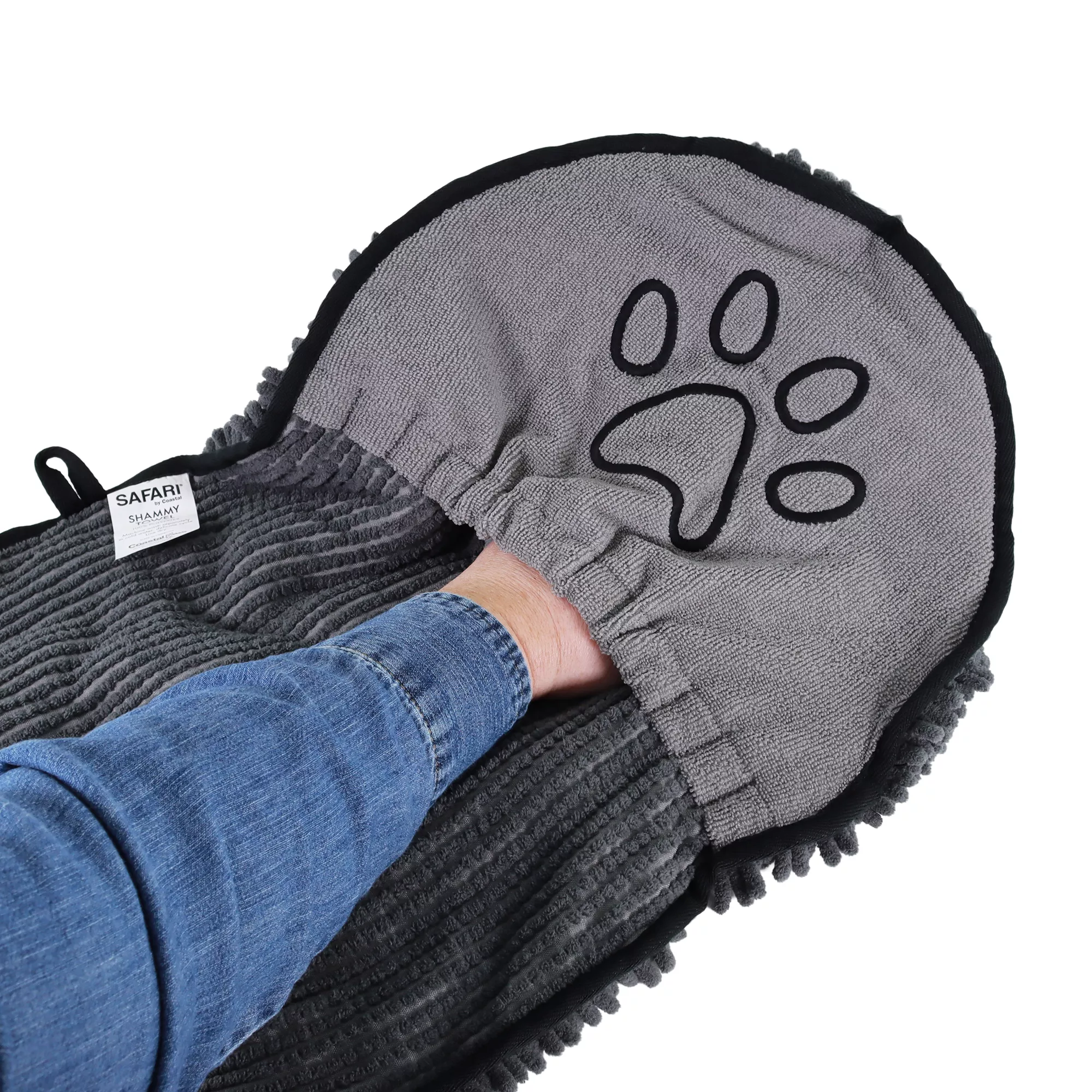 A person holds a gray shammy towel by one of its pocket sections. The pocket has an embroidered black paw‑print design and a black edge trim. The towel’s central panel and opposite pocket are also visible, along with the textured gray microfiber chenille surface at the bottom. The background includes indoor flooring.